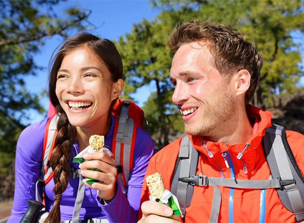 This image shows two non-celebrity hikers taking a snack break in a natural outdoor setting. The image is a widely available stock photo used for various commercial and editorial purposes, such as illustrating articles about healthy snacks or outdoor activities.