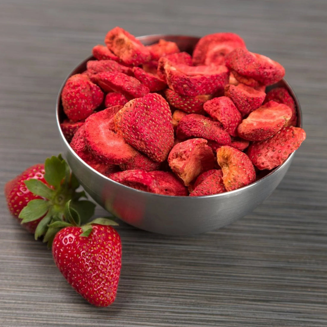 Dried strawberries in a bowl on a wooden surface with fresh strawberries beside it.